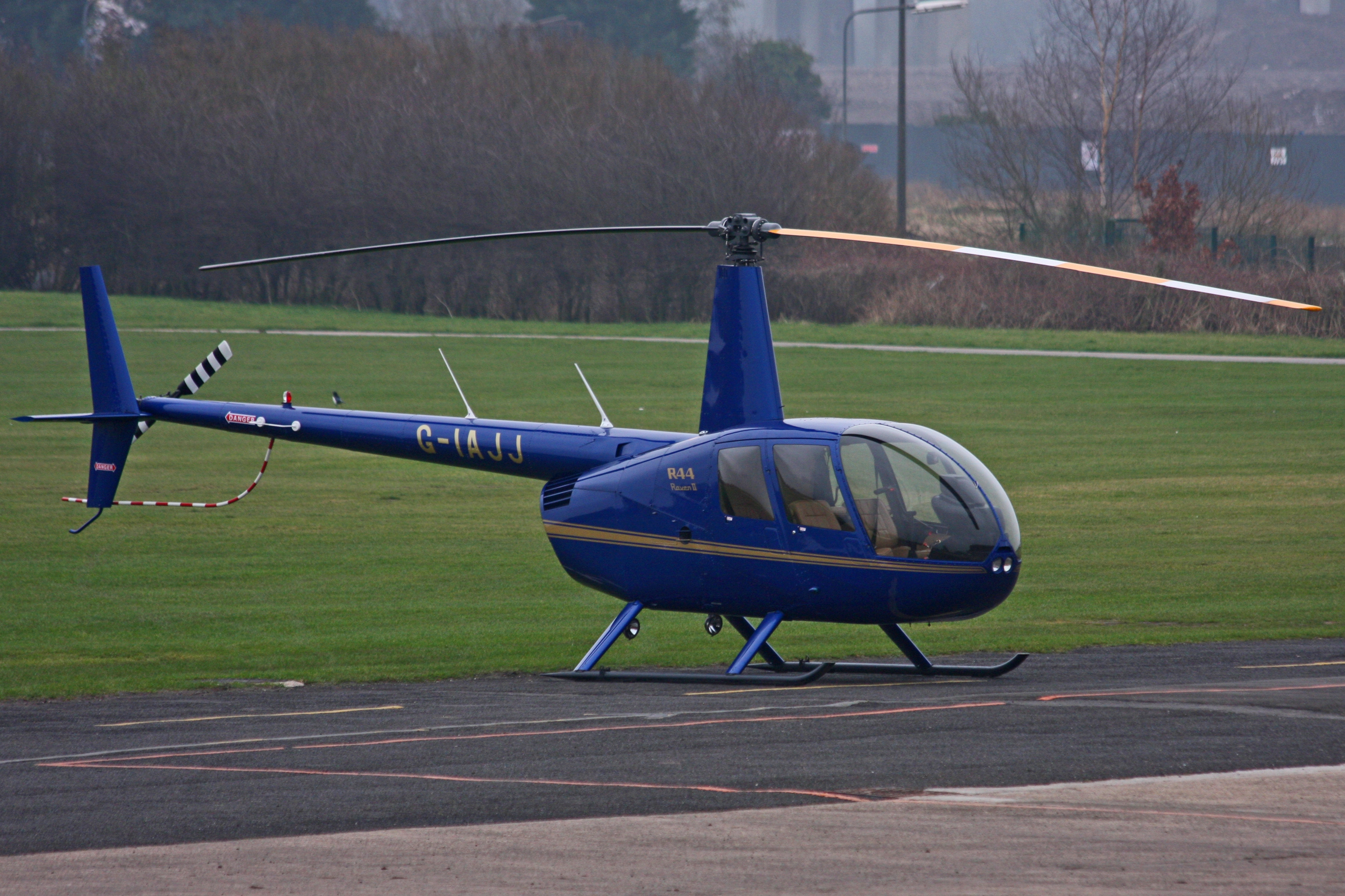 Real blue Robinson R44 helicopter G-IAJJ parked on a paved pad at City Airport Manchester, Barton, with grass and airfield buildings behind.