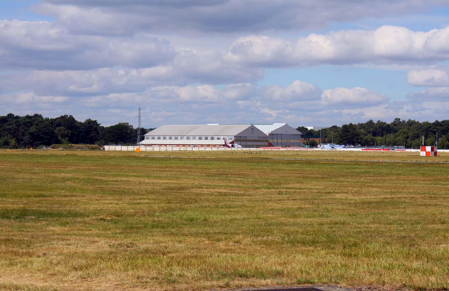 Real photograph across Farnborough Airfield in Hampshire, showing aircraft hangars, business jets on the apron and the wider airfield setting.