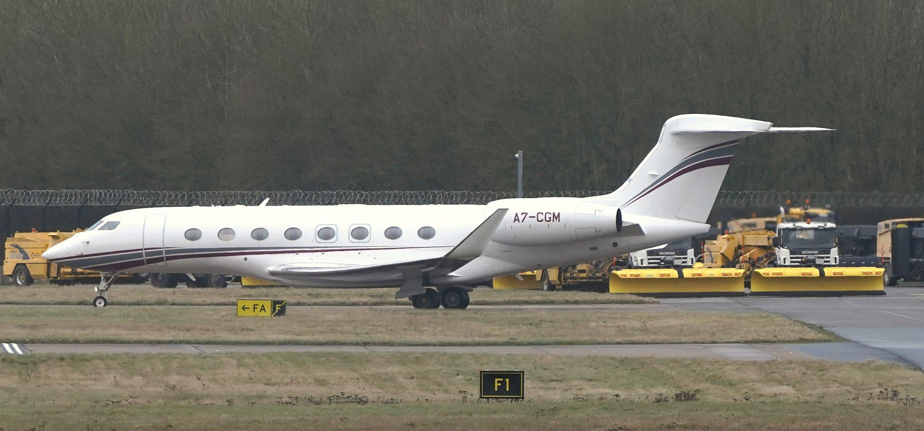 Real photograph of a white Gulfstream G650ER business jet on the airfield at London Stansted, with trees and airport service vehicles behind.