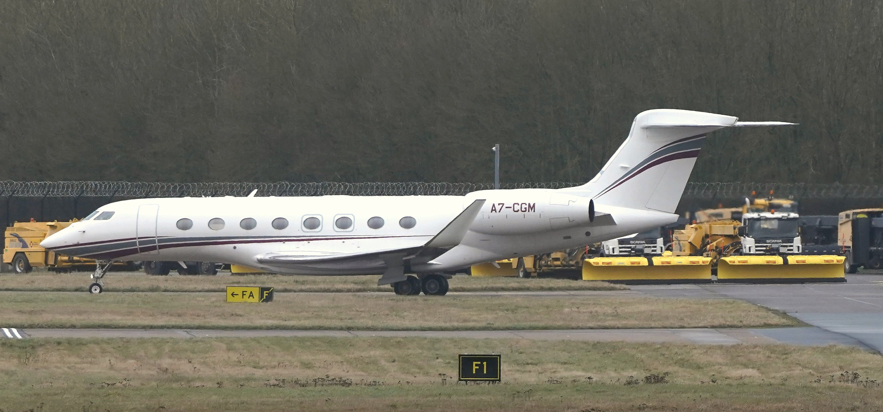 Real photograph of a white Gulfstream G650ER business jet on the airfield at London Stansted, with trees and airport service vehicles behind.