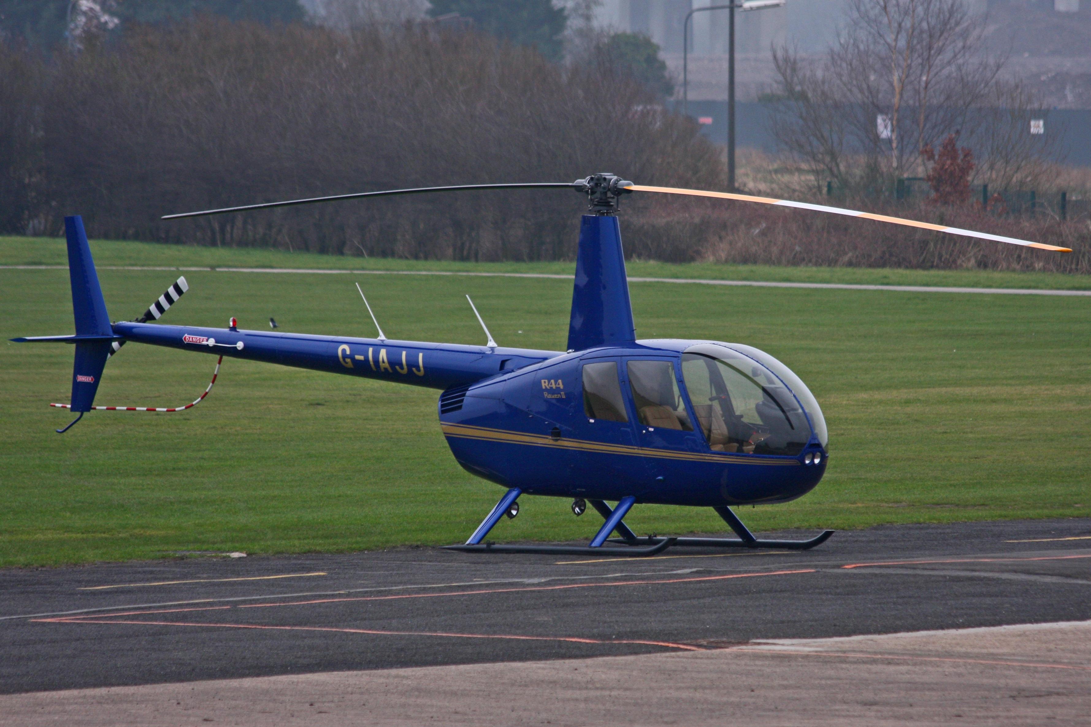 Real photograph of a Robinson R44 helicopter at City Airport Manchester, with grass and airfield buildings behind.