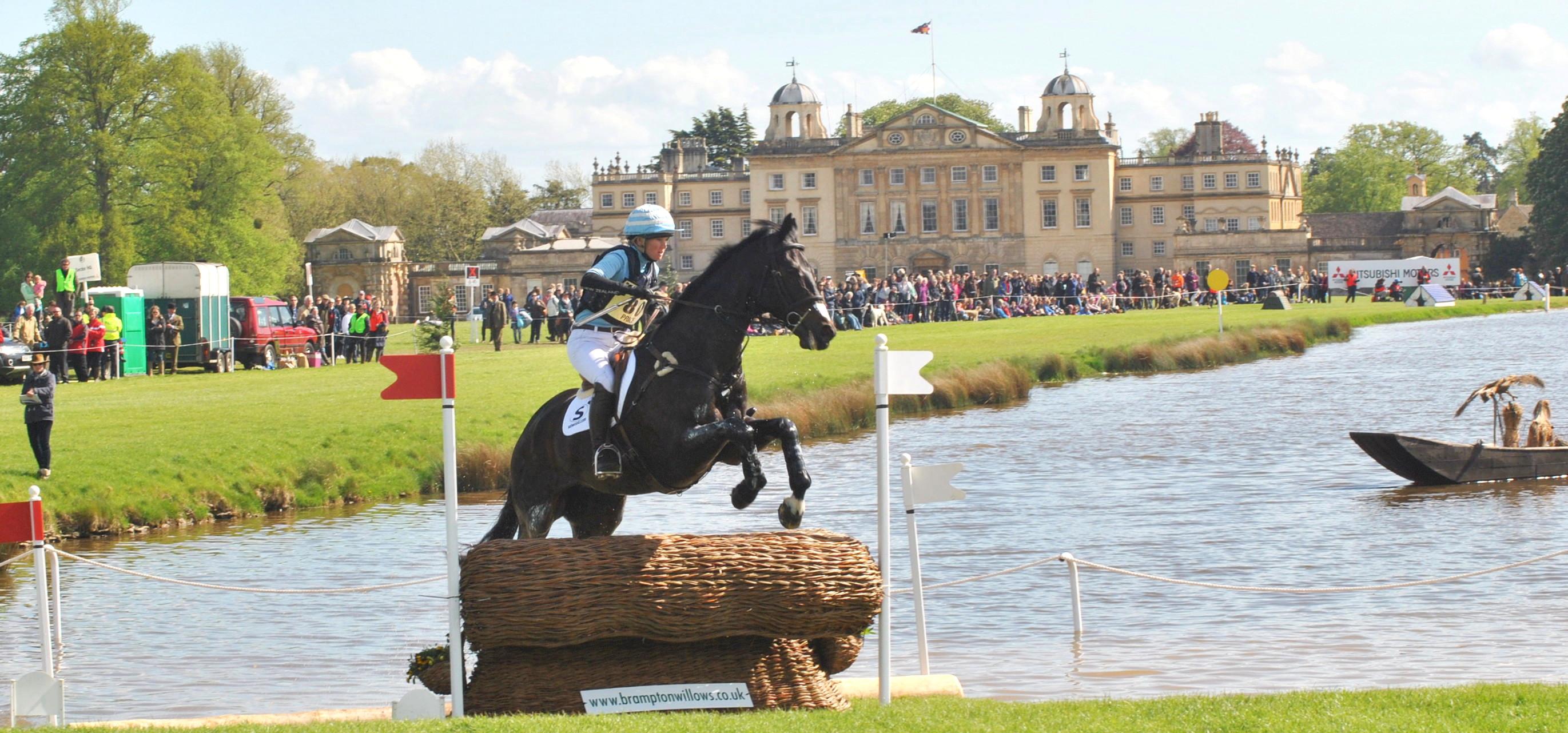 Real photograph of a horse and rider at the water jump during Badminton Horse Trials in Gloucestershire in 2015, with Badminton House and spectators behind.