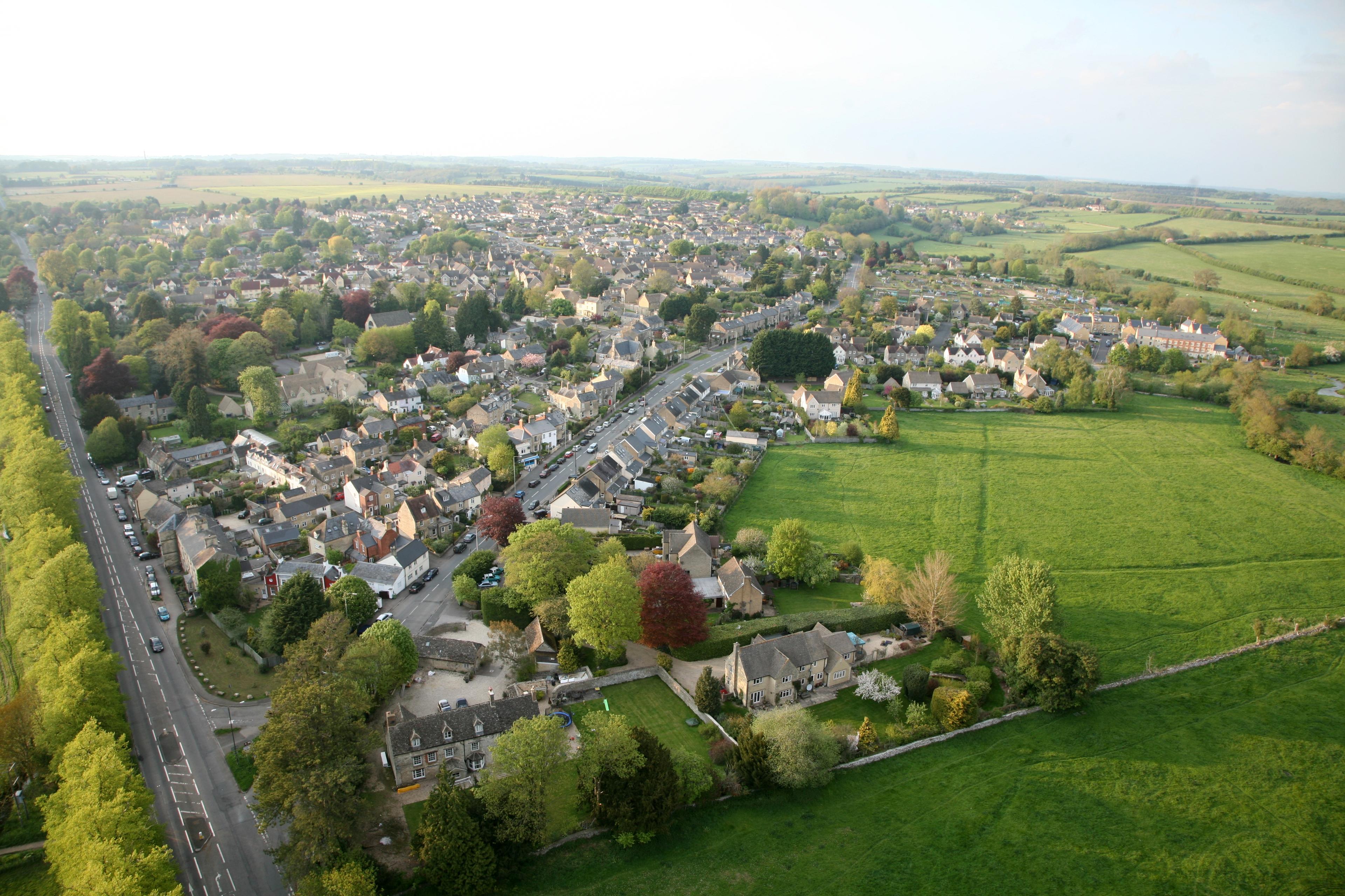 Real aerial photograph of Cirencester, Gloucestershire, showing residential streets, stone-built homes, roads and surrounding agricultural fields at the settlement edge.