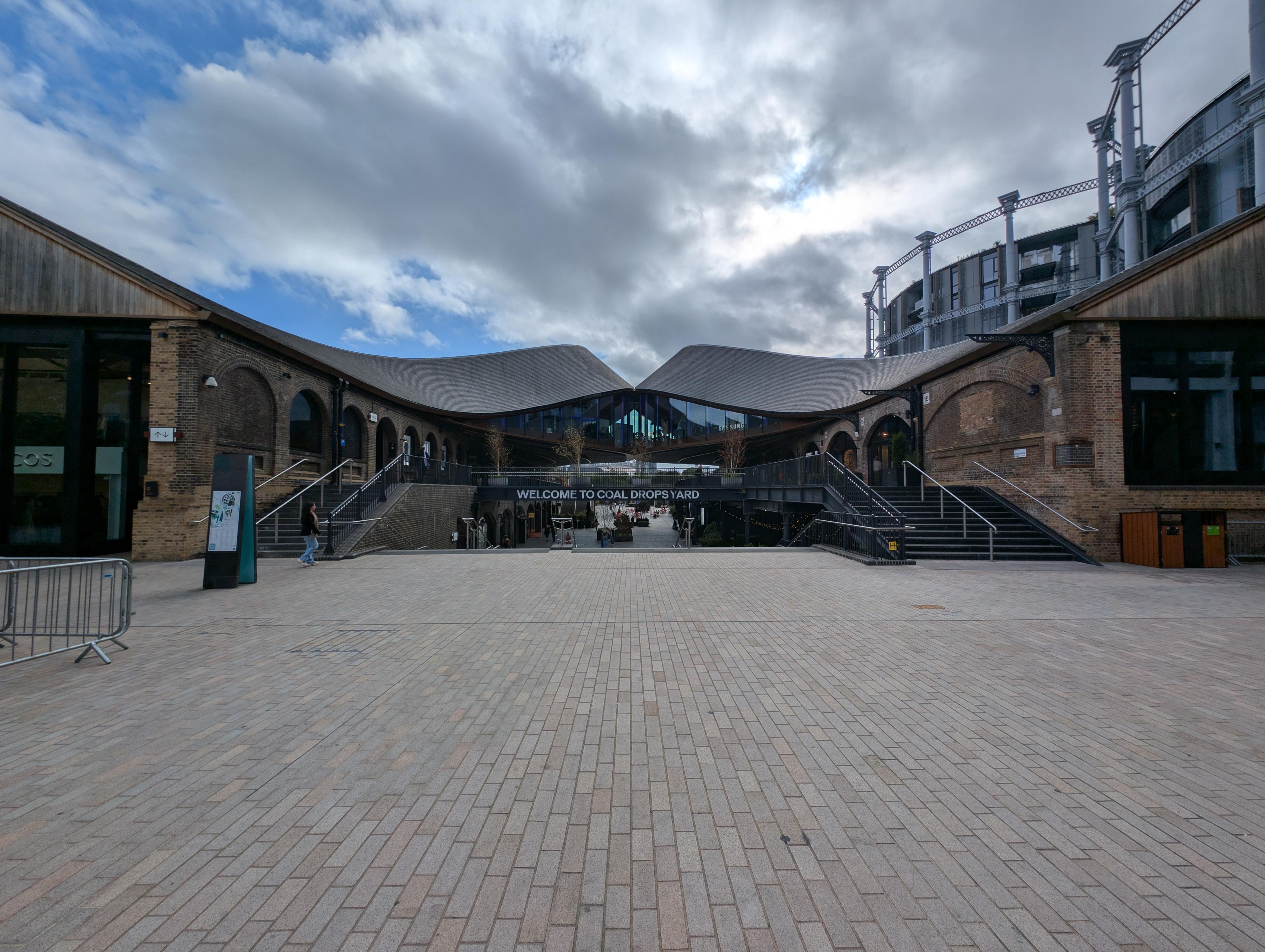 Real view of Coal Drops Yard from the north, showing retained industrial buildings, contemporary roof architecture, retail frontages and the shared public space.