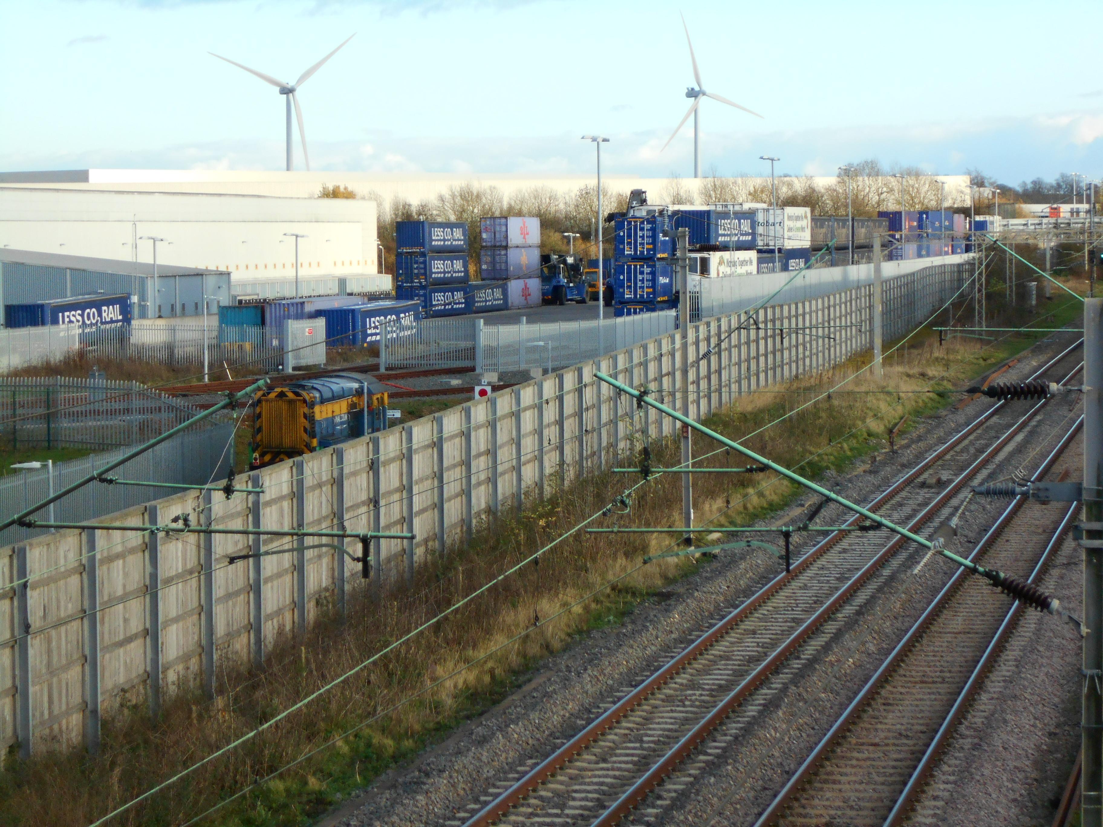Railway lines beside Daventry International Rail Freight Terminal, with a shunting locomotive, stacked freight containers, distribution buildings and wind turbines in the background.