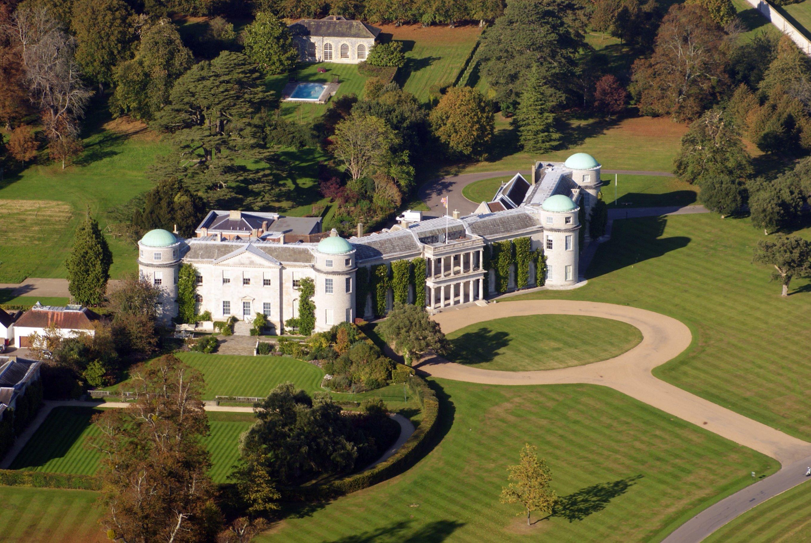 Oblique aerial photograph of Goodwood House, its curved approach drive, estate lawns and mature surrounding trees.