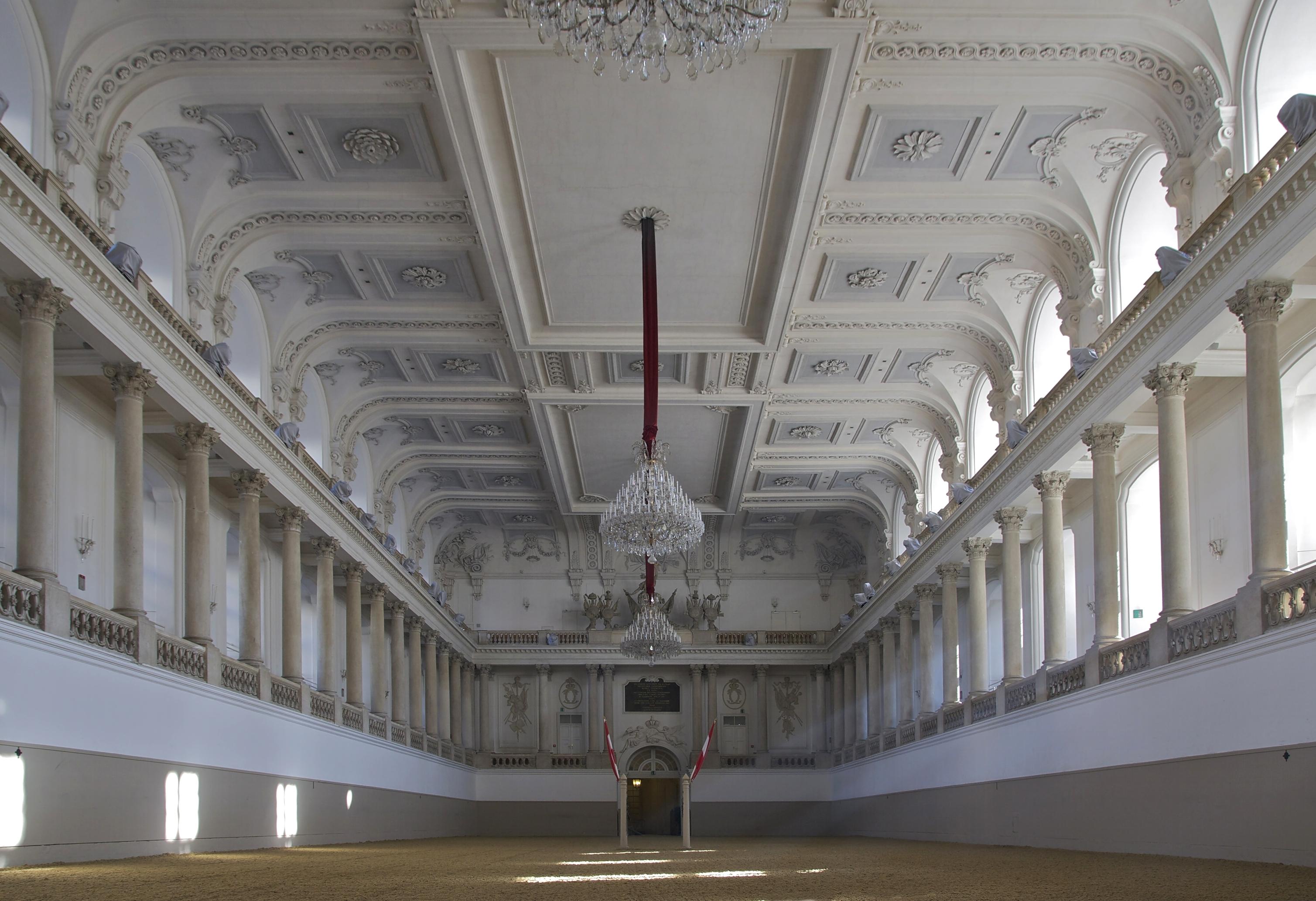 Real photograph of the Spanish Riding School’s winter riding arena in Vienna, Austria, showing its white classical columns, ornate ceiling, chandeliers, galleries and riding surface.
