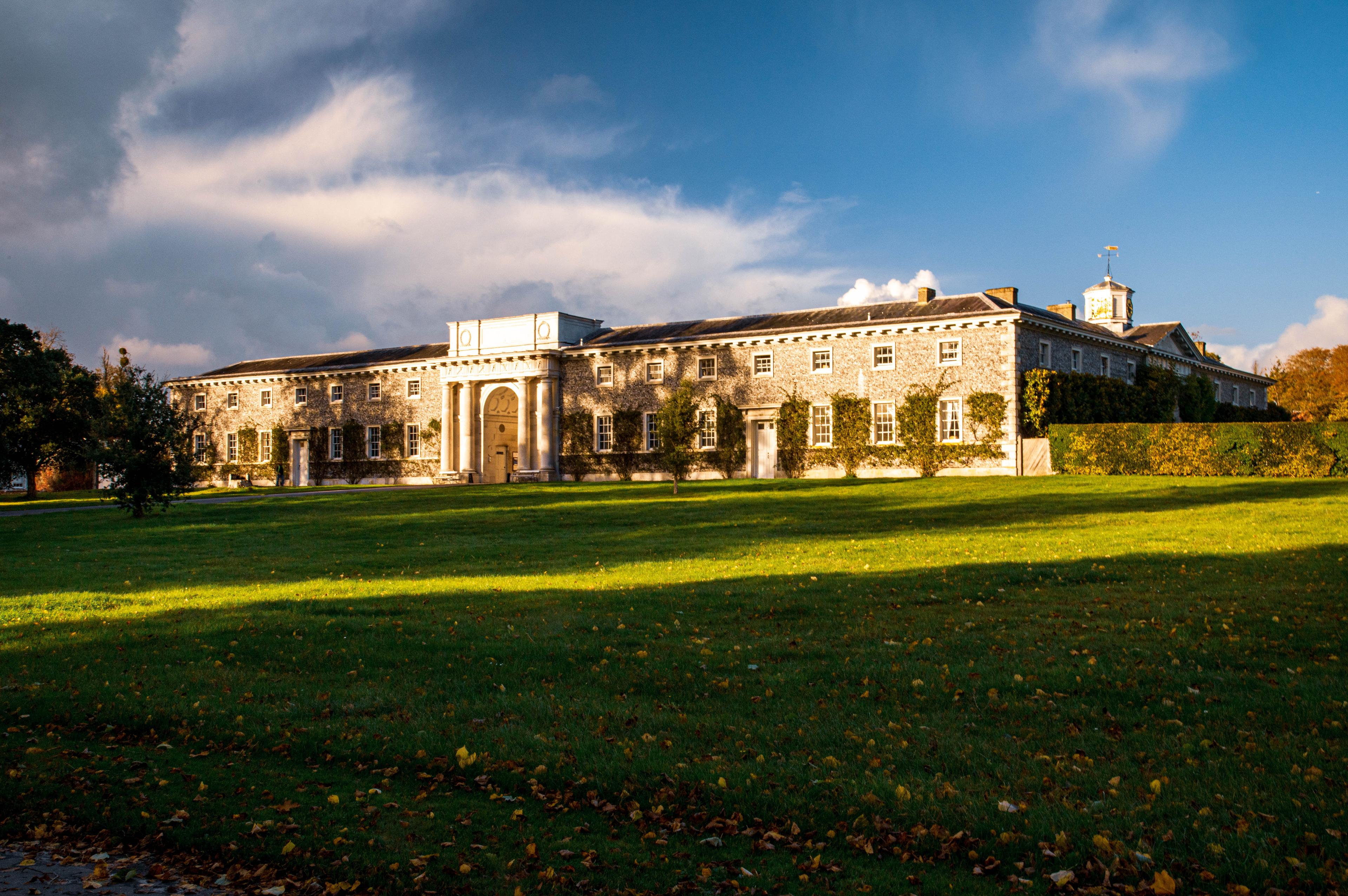 Real photograph of the historic flint stable complex at Goodwood House in West Sussex, showing its long classical frontage, central entrance, clock tower and surrounding estate lawn in late-afternoon light.