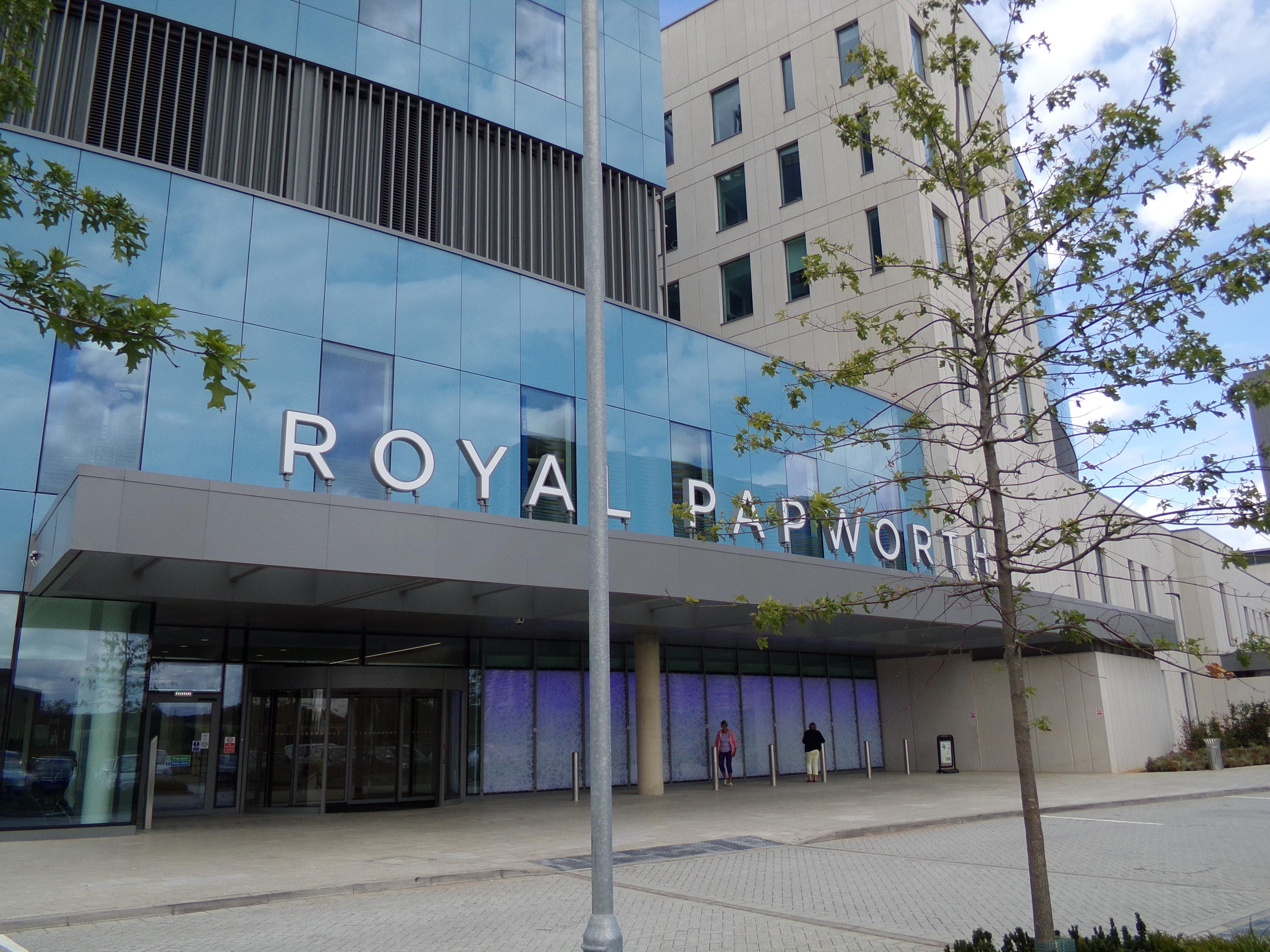 Real photograph of the entrance to Royal Papworth Hospital in Cambridge, showing its glazed facade, landscaped approach and pedestrian access.