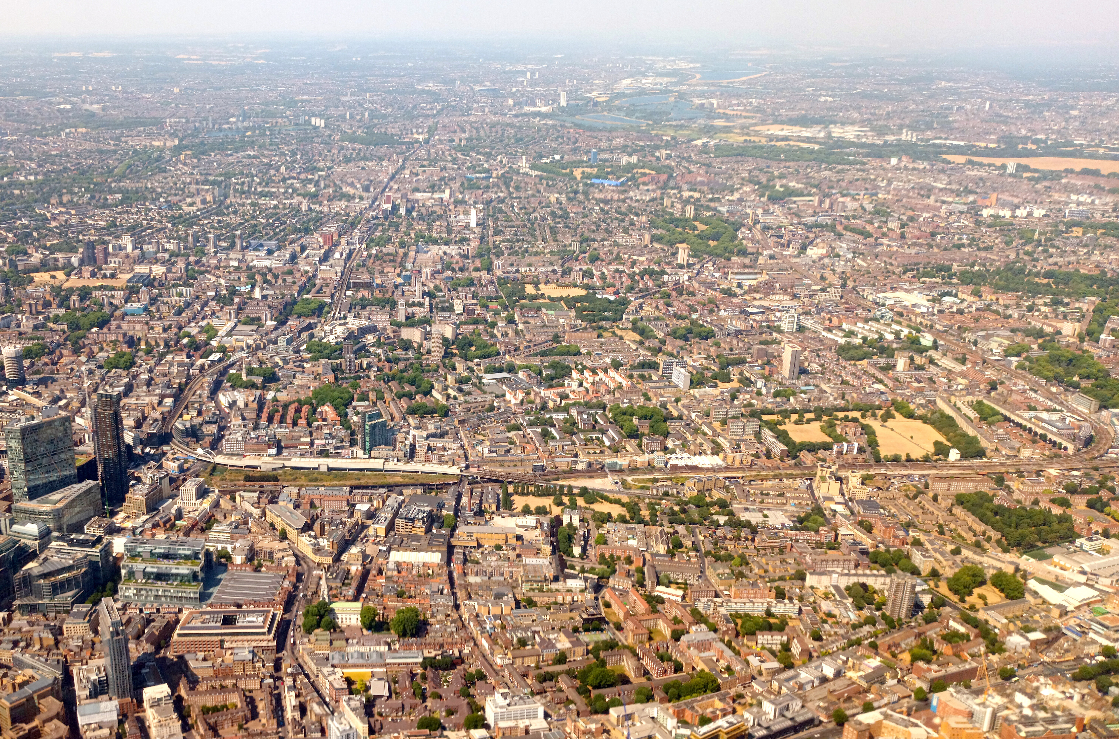 Real aerial photograph of Shoreditch and east London showing commercial buildings, residential neighbourhoods, railway infrastructure and green spaces within the wider urban context.