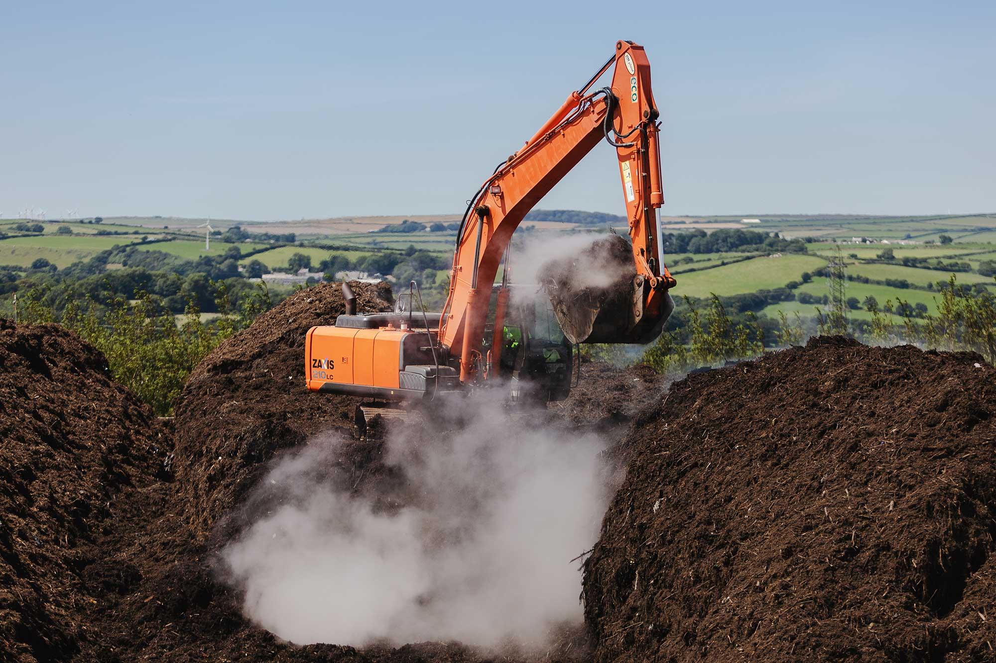 Real photograph from The Green Waste Company showing an excavator handling compost in long rows, with the Cornish agricultural landscape beyond.